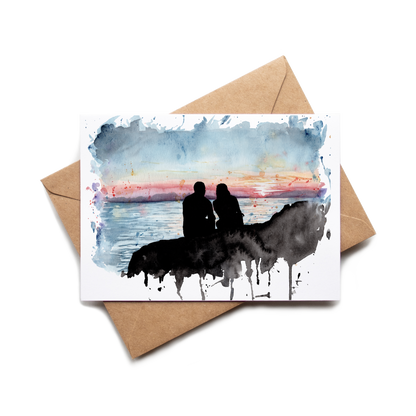 Greeting card with watercolor design of two people on a horse at sunset, placed on a brown envelope.