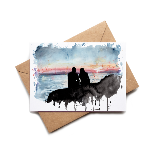 Greeting card with watercolor design of two people on a horse at sunset, placed on a brown envelope.