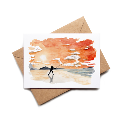 Greeting card with a painting of a person fishing on a beach, placed on a matching brown envelope.