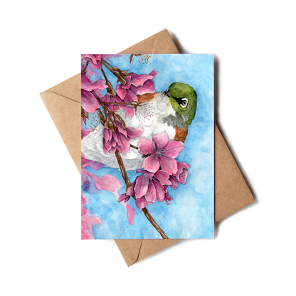 Greeting card with a hummingbird and pink flowers on a blue background, accompanied by a brown envelope.